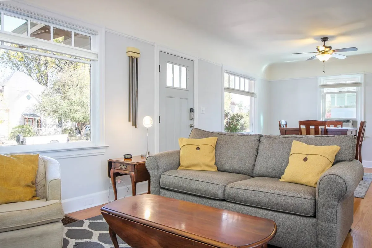 A cozy living room featuring a sofa with cheerful yellow pillows, a wooden coffee table, and a dining area visible in the background. The space enjoys natural light and is styled with warm, inviting decor, perfect for relaxation or social gatherings.