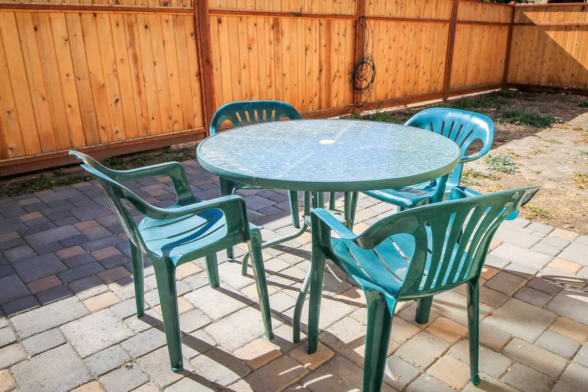The image features a round glass-top table surrounded by plastic chairs on a paved ground, all set within a fenced backyard. The wooden fence provides privacy and a sense of enclosure, while the arrangement invites outdoor dining and social gatherings. The space appears welcoming and relaxed, ideal for both quiet mornings and lively get-togethers.