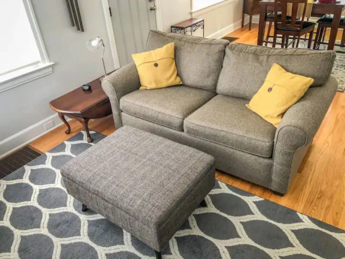 An inviting living space with a sofa, ottoman, rug, and lamp, arranged to encourage relaxation, reading, or socializing. Sunlight filters into the room, enhancing the warm, comfortable atmosphere.