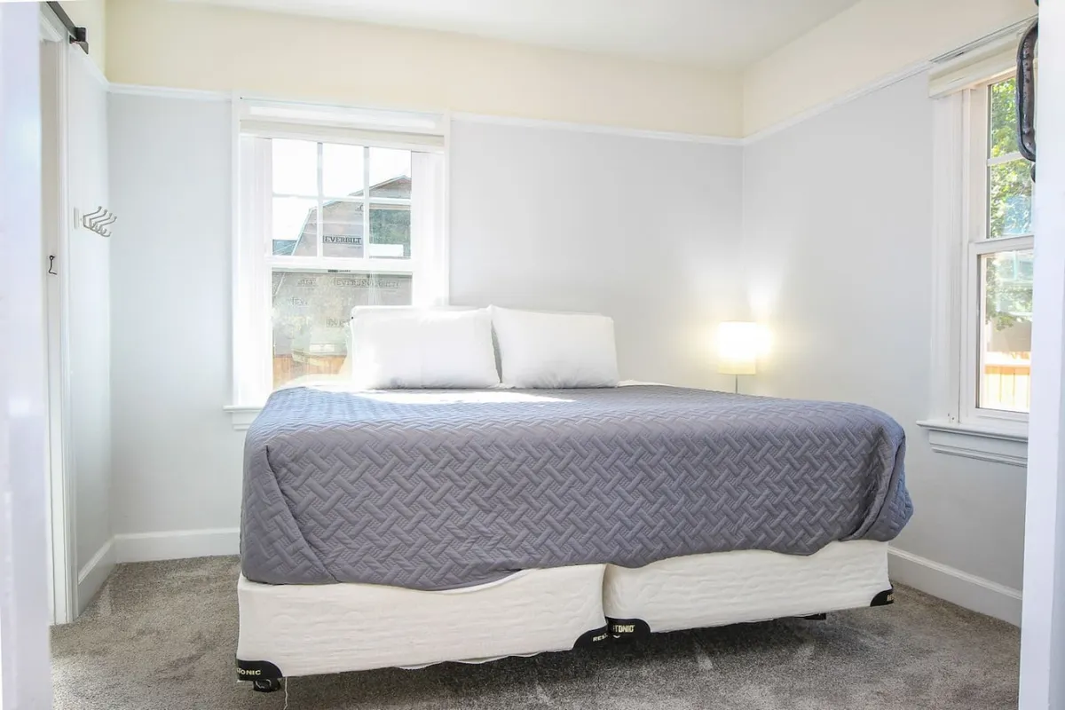 A tranquil bedroom featuring a plush bed with a gray quilt, windows that allow natural light to gently brighten the room, and a thoughtfully placed nightstand with a lamp. The decor is minimalist and soothing, designed to foster relaxation and peaceful sleep.