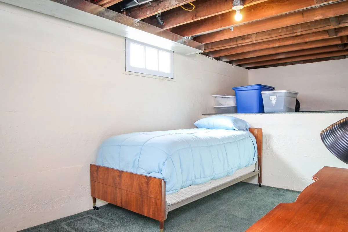 A second bedroom with a neatly made bed, a window bringing in soft daylight, clever storage containers, and a small desk. The space feels calm and functional, suitable for rest or a quiet moment to read or work.
