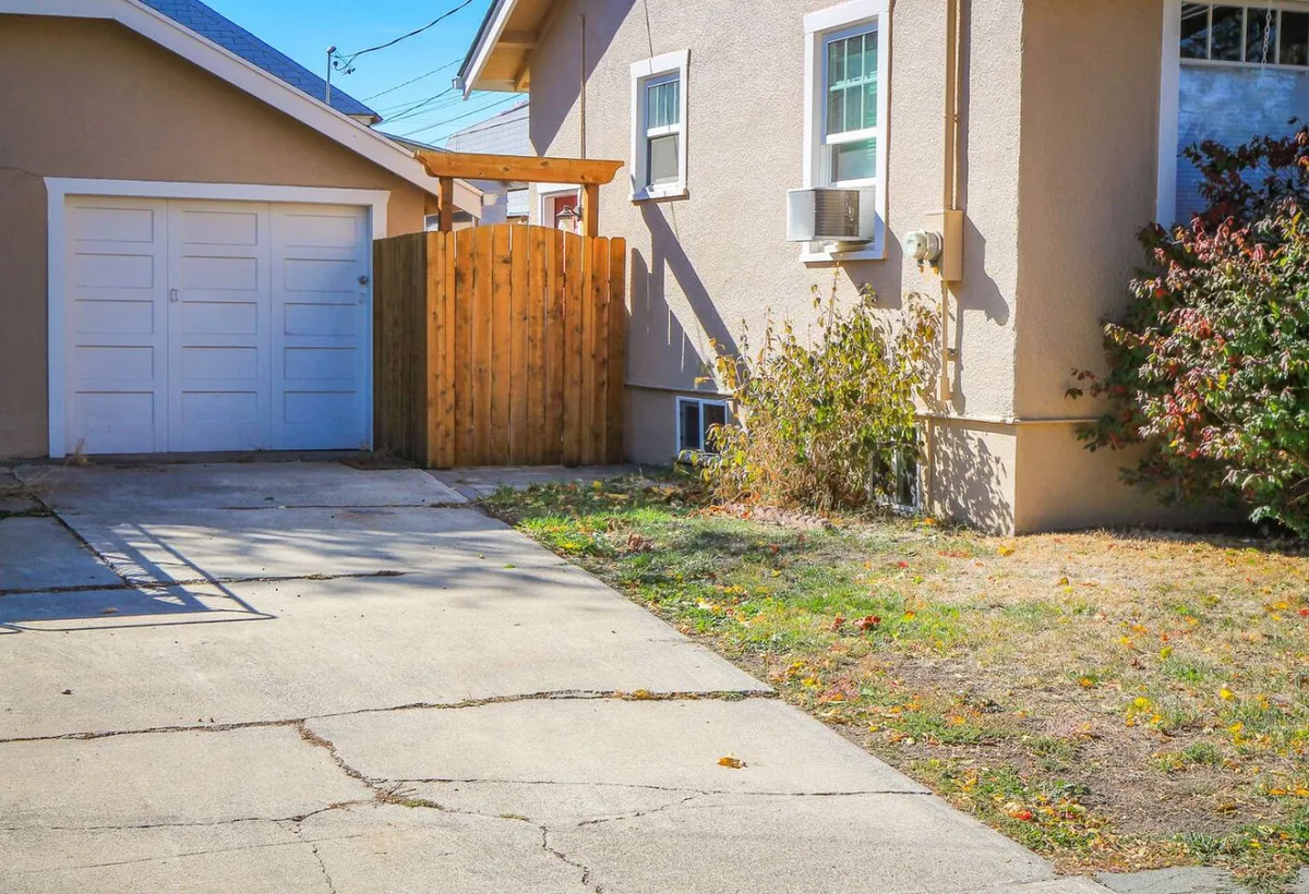 The photo highlights a backyard scene featuring a wooden fence, garage door, air conditioning unit, and a landscaped area with sunlight filtering through trees. The area looks casual and inviting, suitable for various activities such as gardening, children's play, or summer evenings outdoors.