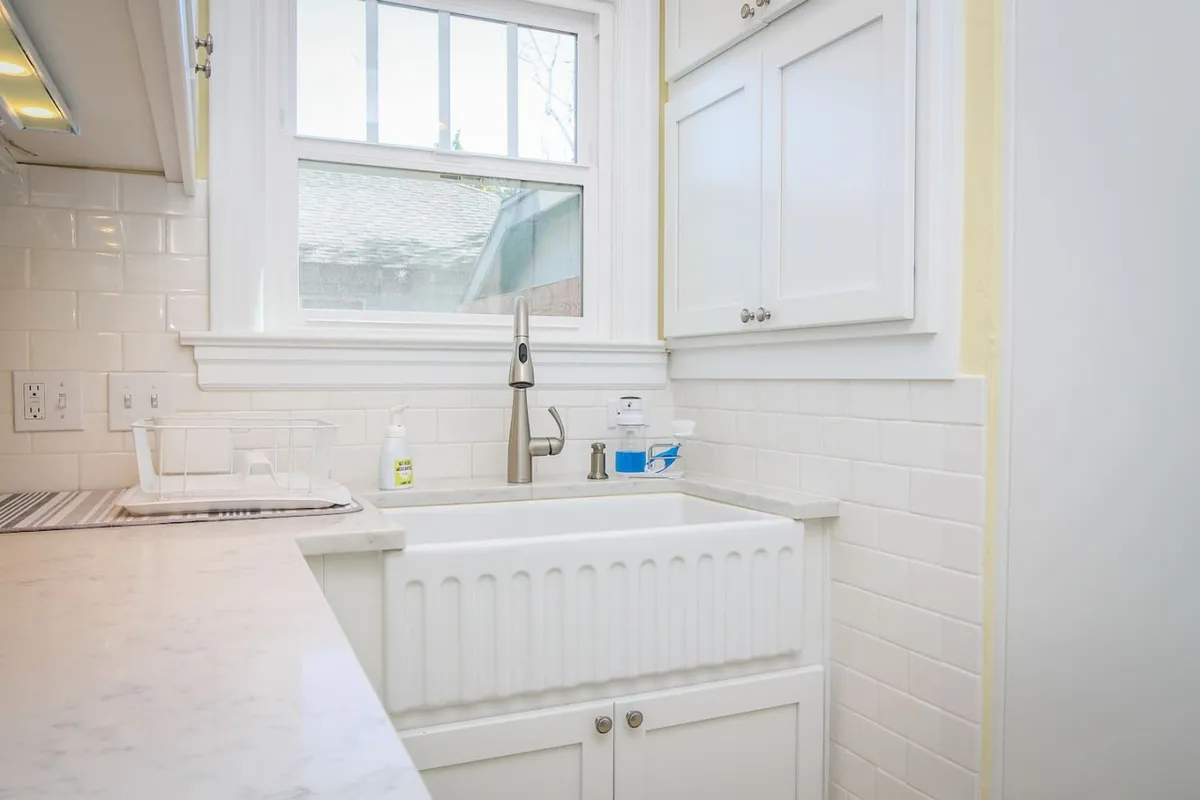 A farmhouse-style kitchen sink with a modern faucet, white cabinetry, and a window streaming in natural light. The setup is clean and functional, ideal for meal prep and socializing while cooking.